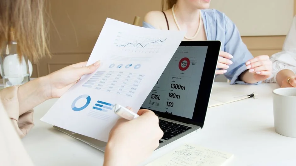 Two businessmen analyze stock market data using a smartphone and laptop in a modern office setting.